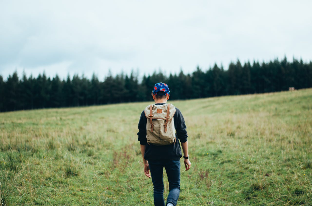 A lone man hiking across a vast, scenic green meadow with forested background.