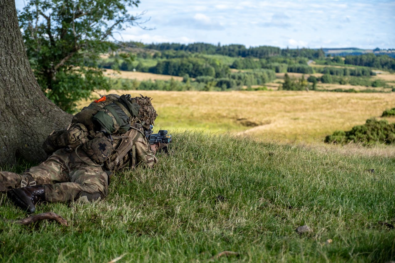 About A soldier in camouflage lies in a rural field during a military training exercise.